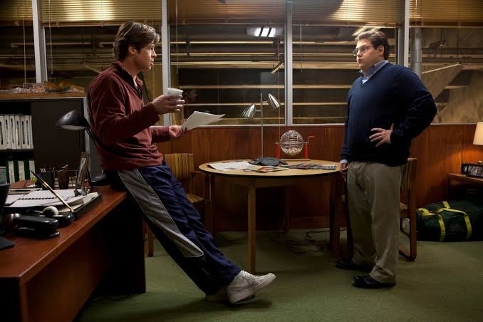 Billy Beane (Brad Pitt) leans against a desk holding papers while Peter Brand (Jonah Hill) stands with hands on hips in the Oakland A's office in Moneyball.
